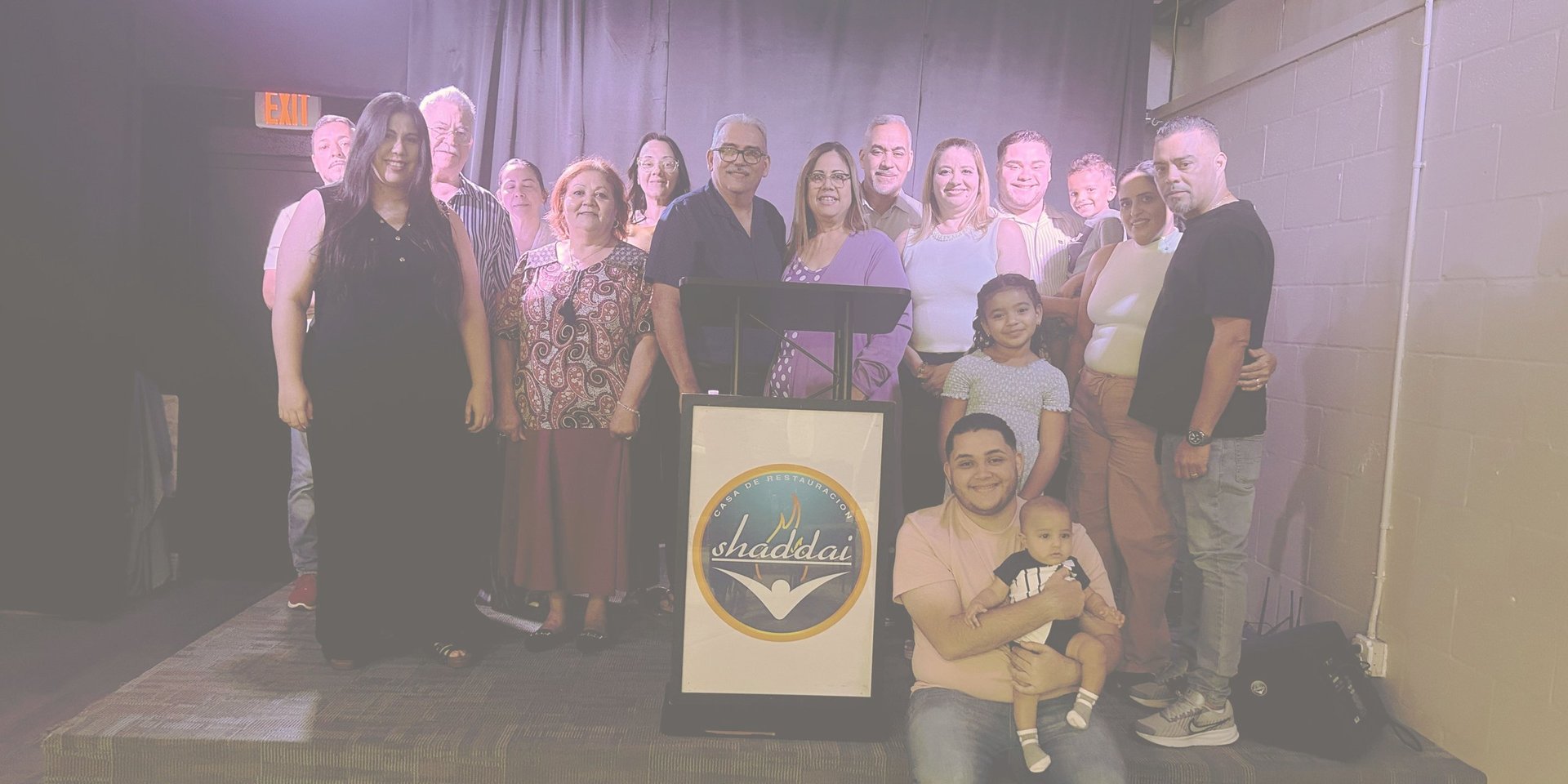 Group of people posing together at an indoor event, standing behind a podium with a logo, with two children in front