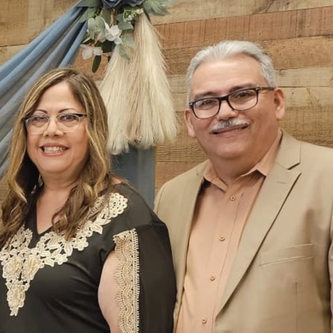 Two adults posing together indoors. Woman on left wearing black dress with lace detail and glasses. Man on right wearing tan blazer and glasses, with white hair and mustache. Wooden interior with holiday decoration visible in background
