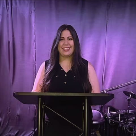 Woman with long dark hair standing at a podium, smiling, with purple lighting and drums visible in the background